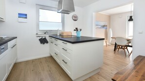 A modern kitchen with white cabinets, a black countertop, and wooden flooring. There is a window with blinds, a clock on the wall, and a blue cup holder with utensils. - NEW 2018 holiday home exclusively near the beach, sauna, fireplace, free WiFi, garden (Wiek /Rügen)