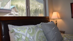 A hotel room with a bed, a nightstand, a lamp, and a window with a view of a mountain and forest.