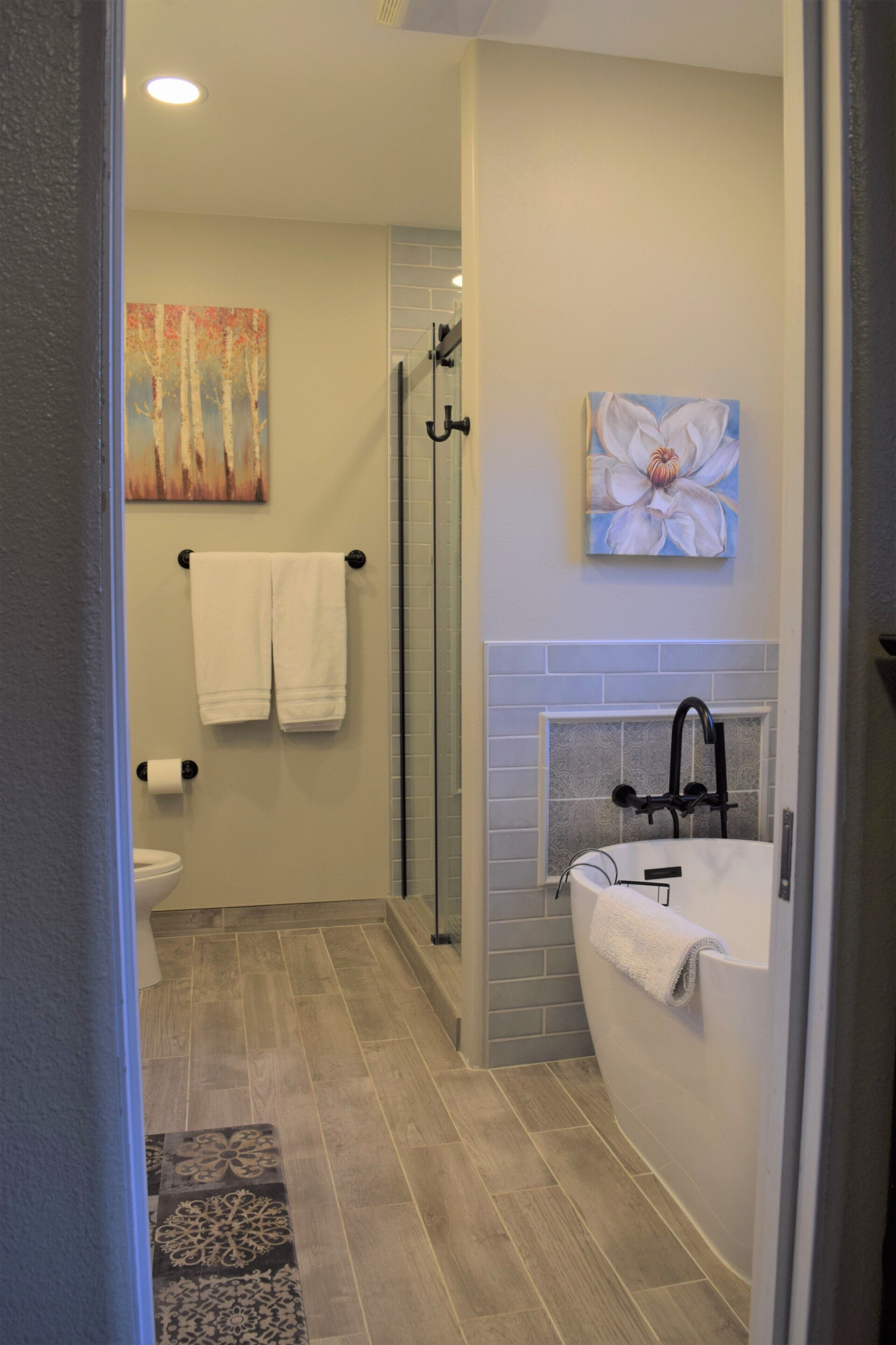 Een badkamer met een douche, toilet en bad. Handdoeken hangen aan de muur en er hangt een schilderij boven het bad.