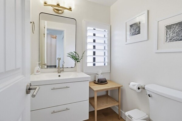 A modern bathroom with a white sink, mirror, and toilet. There is a small wooden table with a tissue box and a plant. - Beach Access, Pool, Spa, Tennis, 2 Bedroom + Private Den (Solana Beach)
