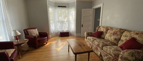 A living room with a wooden floor, a red armchair, a floral sofa, and a small table with a lamp.
