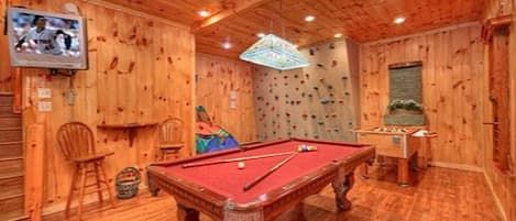 A wooden room with a pool table, chairs, a television, and a climbing wall.