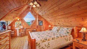 Une chambre confortable située dans les combles, avec un décor intérieur en bois, un lit recouvert d’une courtepointe, un bureau, une chaise et une vue sur le ciel grâce à une lucarne.