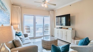 A living room with a sofa, a round table, a television, and a balcony with outdoor furniture.