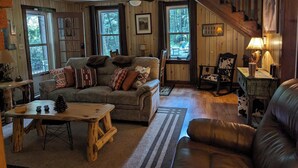 A cozy cabin interior with a wooden floor, a sofa with patterned cushions, a coffee table, a chair with a cow print cushion, and a staircase. - Montgomery Run cabin located in a secluded wooded area close to Clearfield, PA (Clearfield)