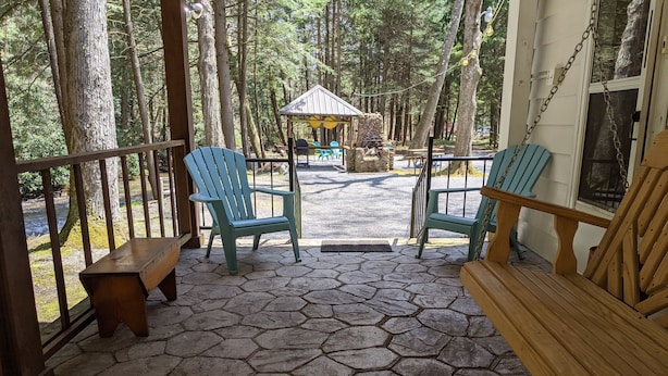 A covered outdoor seating area with hexagonal stone flooring, two light blue Adirondack chairs, a wooden bench, and a small table. - Montgomery Run cabin located in a secluded wooded area close to Clearfield, PA (Clearfield)