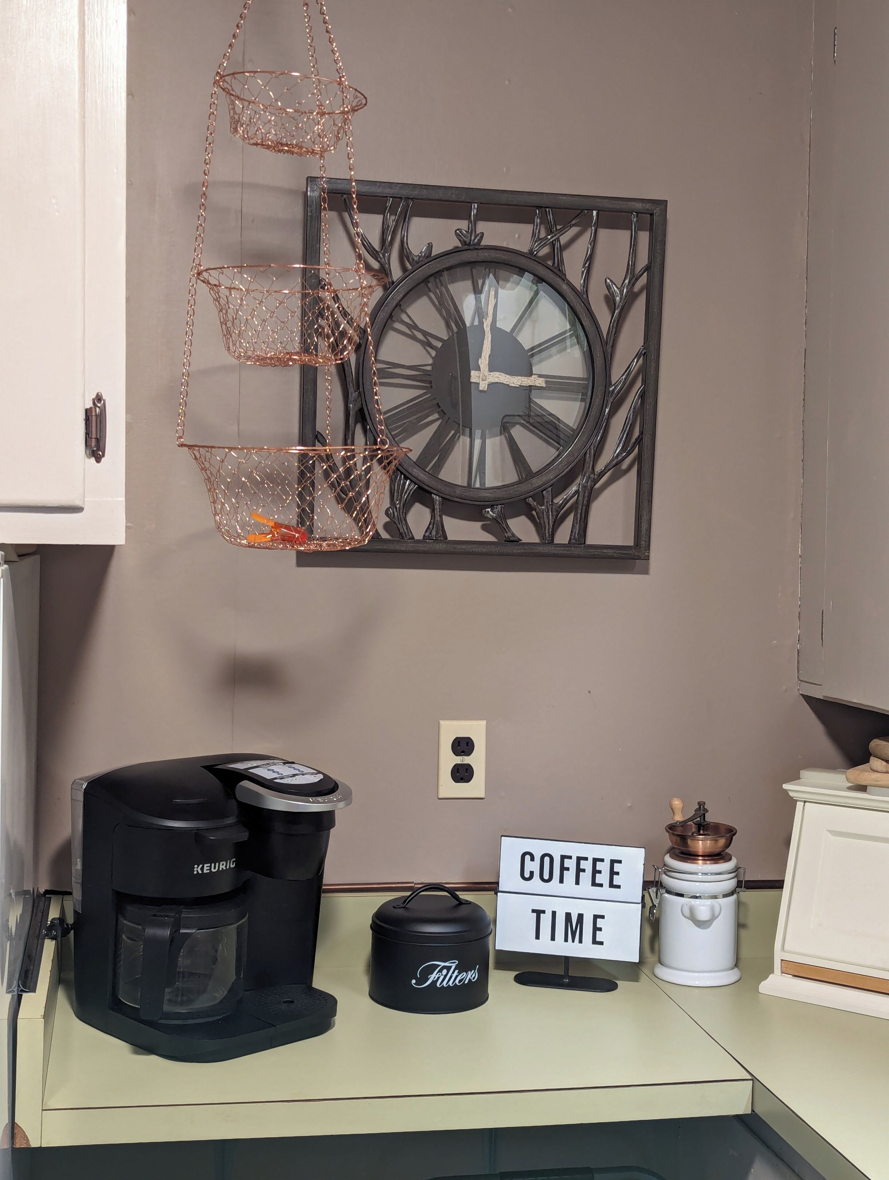 Une petite cuisine équipée d’une machine à café, d’une pancarte indiquant « PAUSE CAFÉ », d’une horloge et d’un élément décoratif en métal.