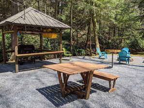 Ein hölzerner Pavillon mit Grill, Picknicktisch und blauen Adirondack-Stühlen inmitten bewaldeter Landschaft.