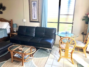 Un salon avec un canapé en cuir noir, une table basse en verre, des fauteuils en osier, une petite table ronde et un tableau encadré accroché au mur.