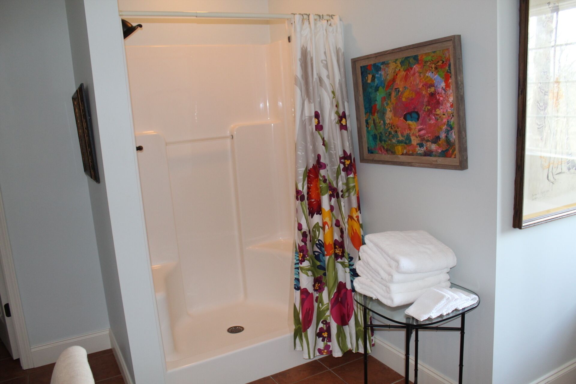 Une douche avec un rideau à motifs floraux, une table en verre avec des serviettes et un tableau accroché au mur.