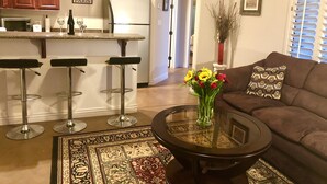 A modern kitchen with wooden cabinets, a white refrigerator, and a bar area with stools. There is a dining area with a round table and a sofa with patterned cushions. - Casita Del sol, 1 BR 1BA-Sleeps 2. Minimum Stay 4 nights. Great North Phoenix (Phoenix)