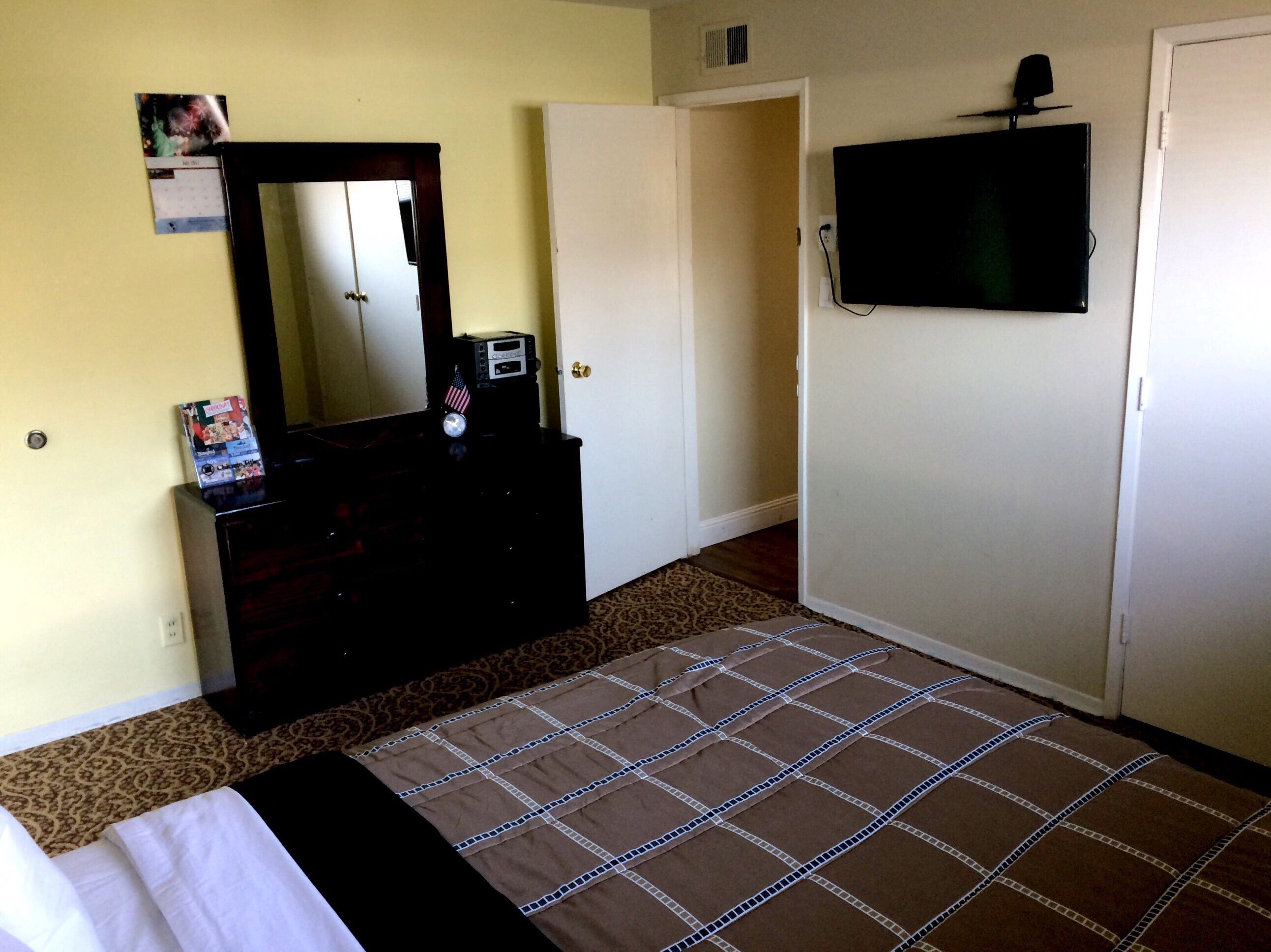 A hotel room with a bed, a dresser with a mirror, a television mounted on the wall, and a door.