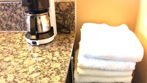 A kitchen with a coffee maker, a granite countertop, a towel rack with folded towels, and a small metal shelf with toiletries.