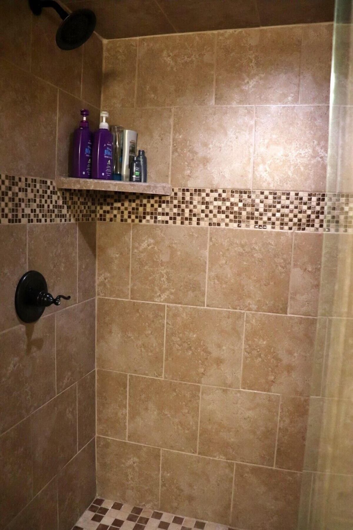 A shower area with beige tiles, a shelf with toiletries, and a showerhead.
