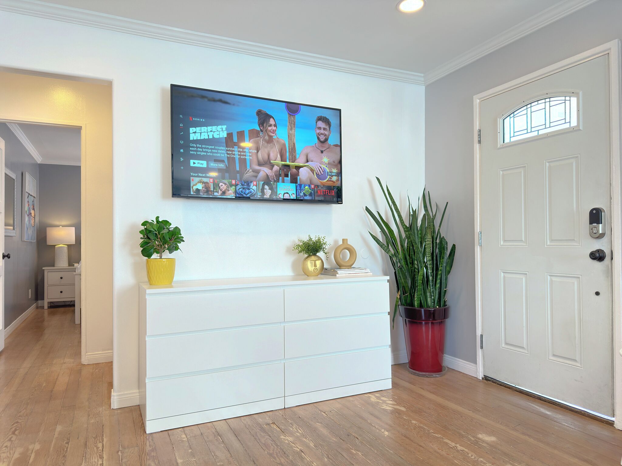 Un saló amb un televisor de pantalla plana muntat a la paret, una taula blanca de consola amb objectes decoratius i una planta en test.