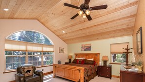 A bedroom with a large bed, a wooden ceiling fan, a window with blinds, a chair with a bear pillow, and a nightstand with lamps. - Charming Dog-Friendly Lake Tahoe Cabin – Hot Tub, Pool Table & Walk to Beach (Carnelian Bay)