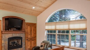 A cozy room with a stone fireplace, a television mounted above it, a comfortable chair with a decorative pillow, and a large window with blinds. - Charming Dog-Friendly Lake Tahoe Cabin – Hot Tub, Pool Table & Walk to Beach (Carnelian Bay)