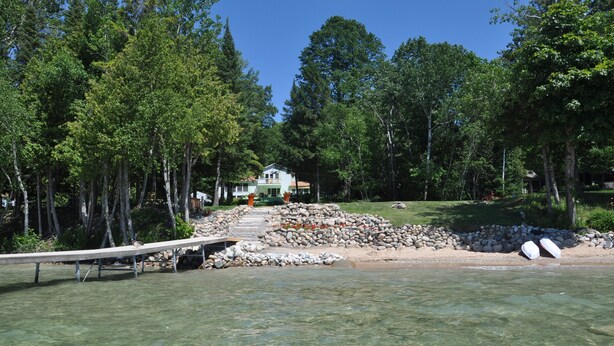 A lakeside property with a dock, clear water, and a house surrounded by trees. - Sunsets,140' on Torch Lk,Lg Dock,Hot Tub,Tennis/Pickle Crt,Kayaks,Play/swing set (Bellaire)