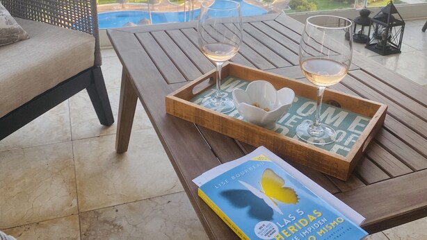 Un balcon avec vue sur une plage et une piscine, une chaise en osier, une table en bois sur laquelle se trouve un plateau avec des verres et un livre intitulé « Lise Bourbeau ».
