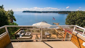 A deck with outdoor furniture, a flag, and a view of a lake and distant land. - Anderson Island Luxury Beach Home. Kayak, HotTub, Paddle Board, Beachfront, View (Anderson Island)