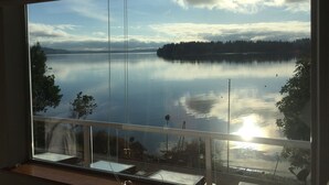 A large window with a view of a calm lake, surrounded by trees and a distant shoreline. - Anderson Island Luxury Beach Home. Kayak, HotTub, Paddle Board, Beachfront, View (Anderson Island)