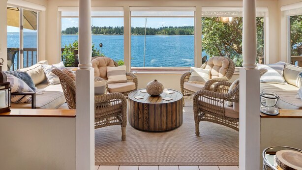 A sunlit room with wicker furniture, large windows, and a view of a lake. - Anderson Island Luxury Beach Home. Kayak, HotTub, Paddle Board, Beachfront, View (Anderson Island)