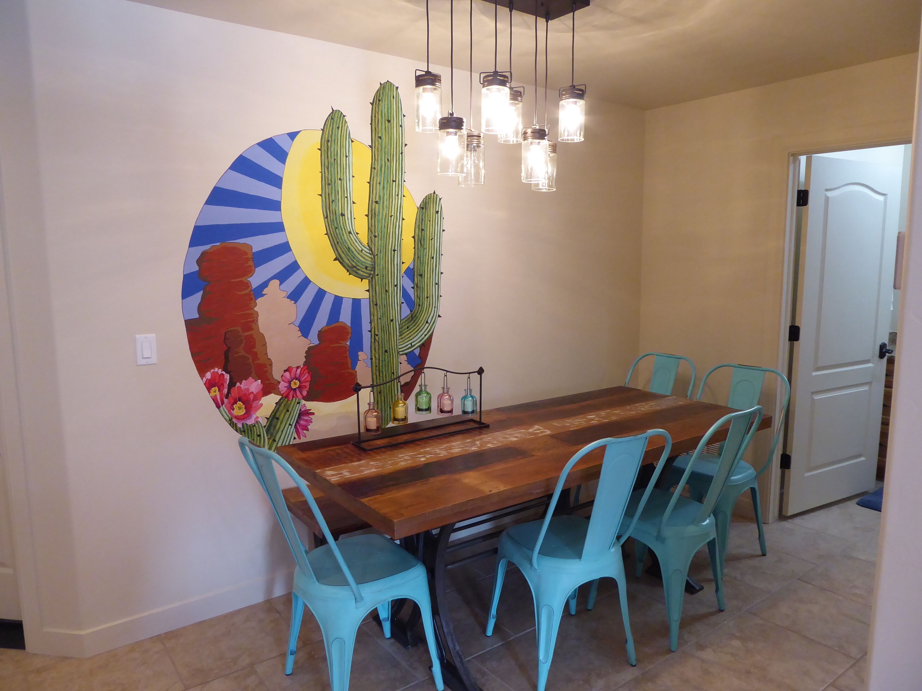 Een eethoek met een houten tafel, turquoise stoelen, een cactus muurschildering en hangende lampen.