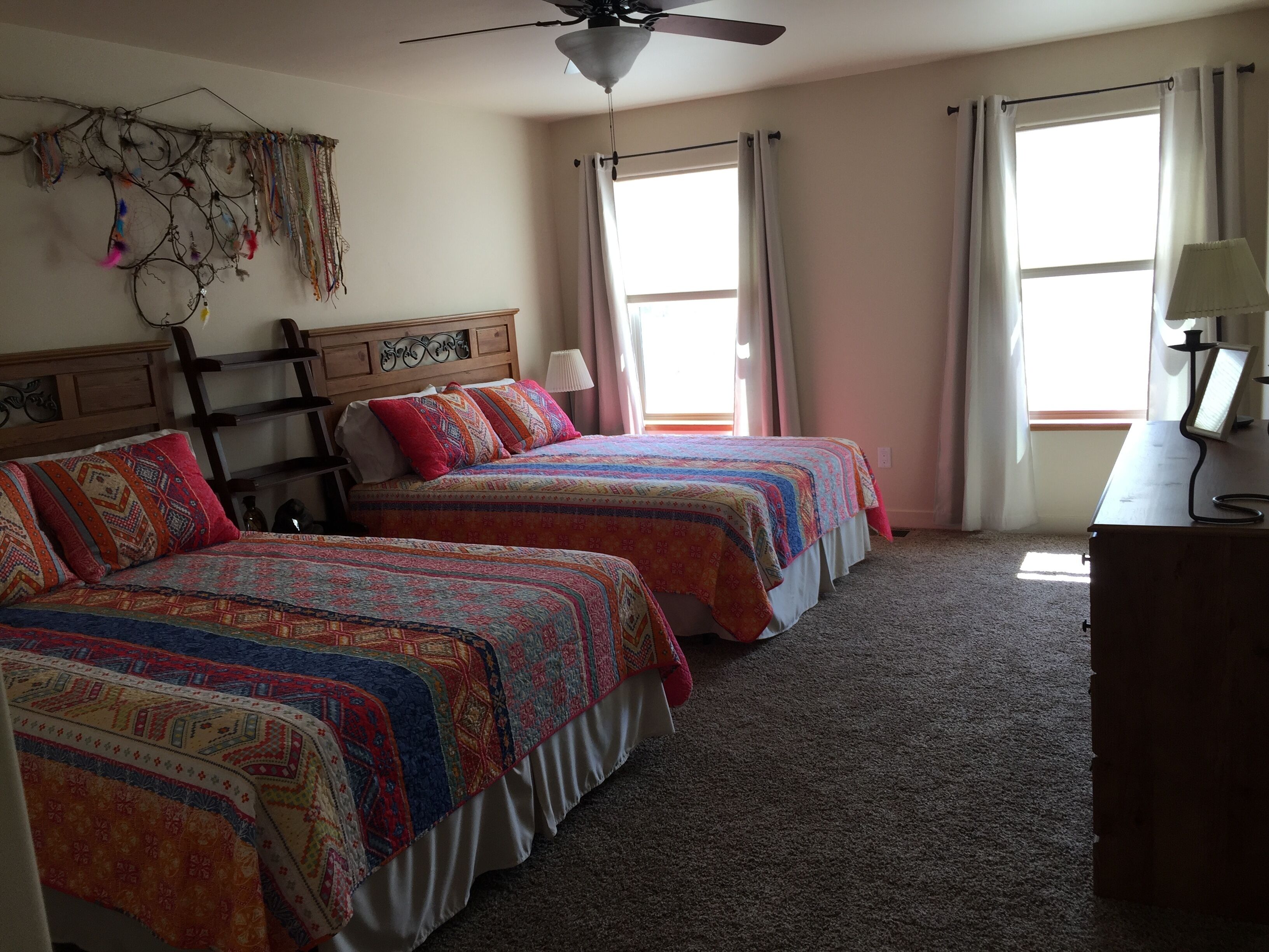 A bedroom with two beds, a ceiling fan, a desk with a lamp, and a window with curtains.