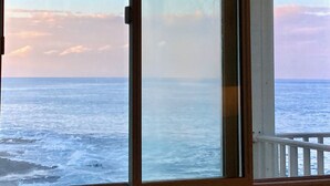 A view from a room with a large sliding glass door to the ocean, featuring a wooden ceiling and a white railing.