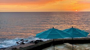 A sunset view from a seaside terrace with two blue umbrellas and a stone balustrade.