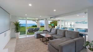 A modern living room with a large grey sectional sofa, a small wooden coffee table, and a large window offering a view of the sea.