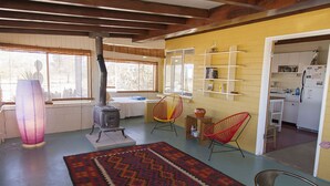 A cozy living room with a wood-burning stove, a patterned rug, and a kitchen area with shelves and a refrigerator. - Midcentury Desert Escape (Twentynine Palms)