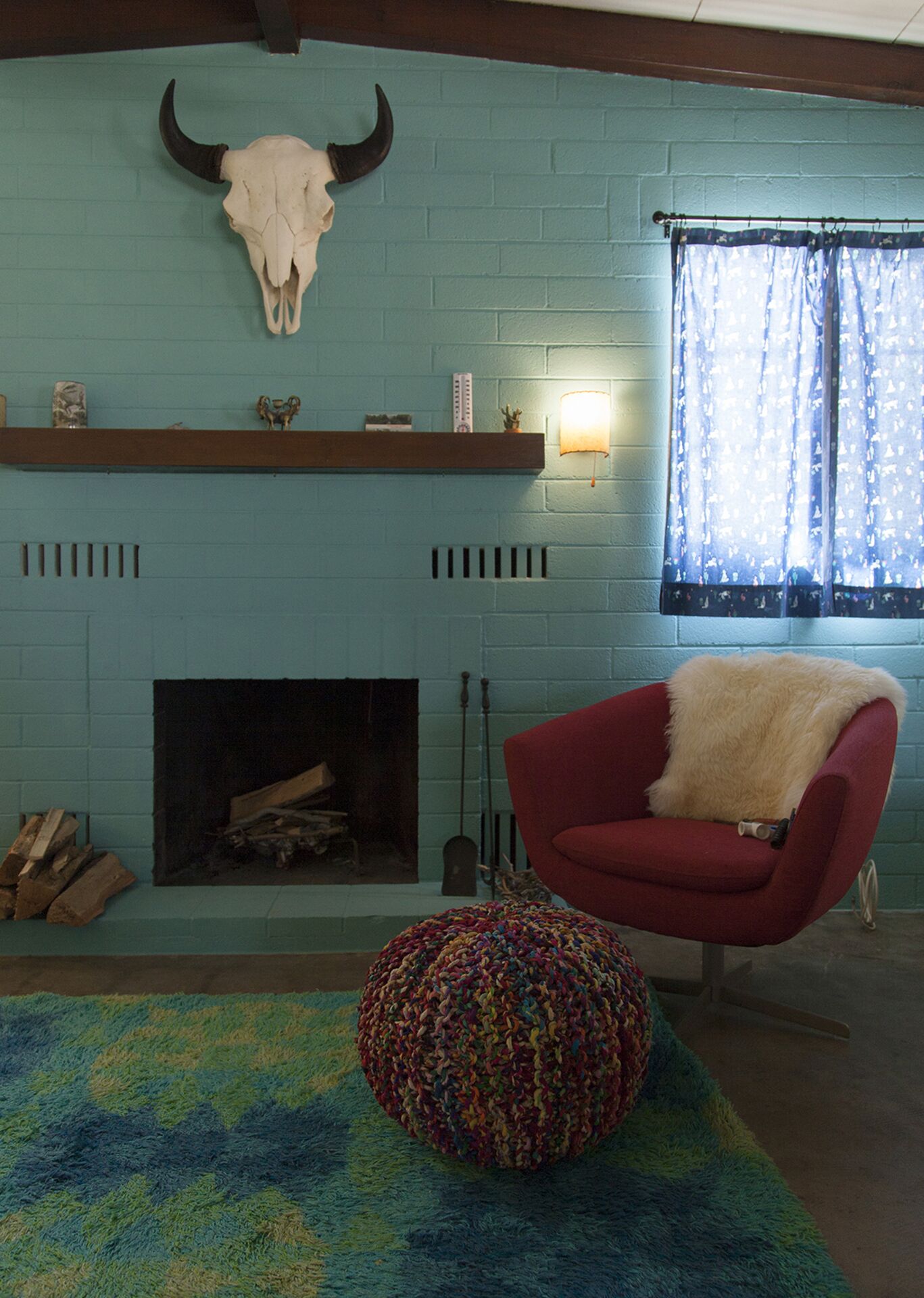 Um quarto com lareira de tijolos azuis, uma poltrona vermelha com manta de pele, um banquinho de tricô colorido e um crânio de vaca fixado na parede.