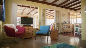 A living room with a red chair, a blue armchair, a television, and a kitchen area with a table and chairs. - Midcentury Desert Escape (Twentynine Palms)
