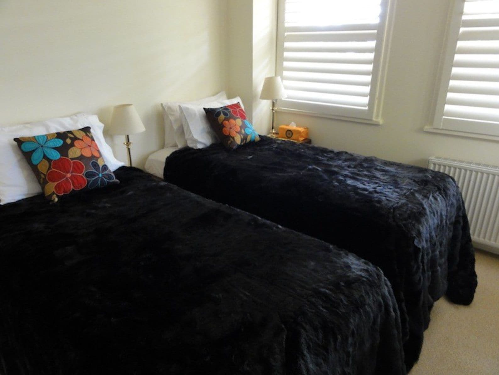 A room with two black beds, white pillows, and a colorful flower-patterned pillow. There is a lamp on each bed and a radiator on the wall.
