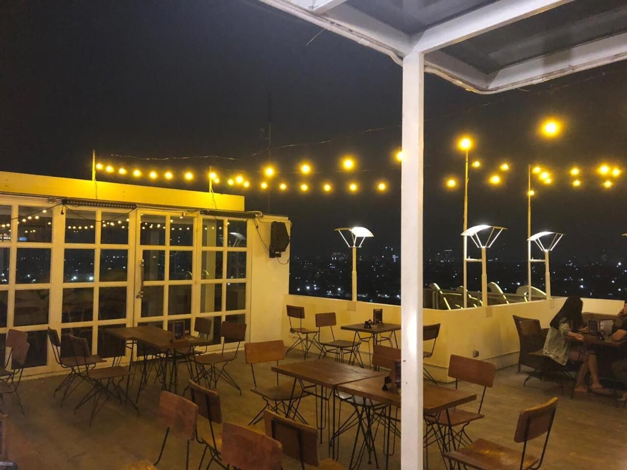 An outdoor terrace with tables and chairs, string lights, and a view of the city at night.