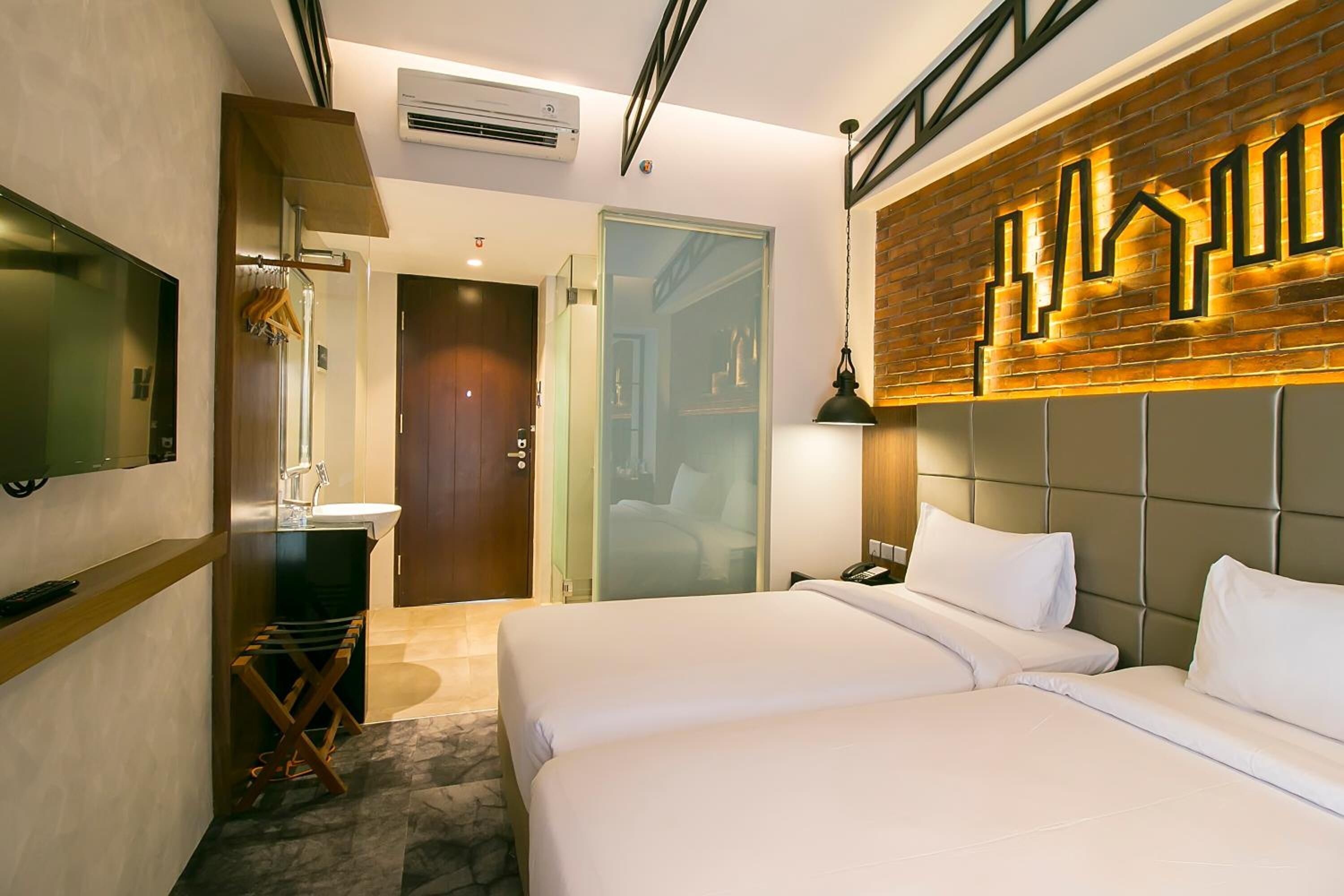 Habitación de hotel con dos camas, un espejo grande, un televisor de pantalla plana, una mesita de noche de madera y una pared con diseño de ladrillo.