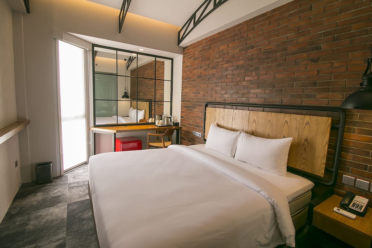 Een hotelkamer met een groot bed, een bureau met stoel, een rode prullenbak, een houten hoofdbord en een bakstenen muur.
