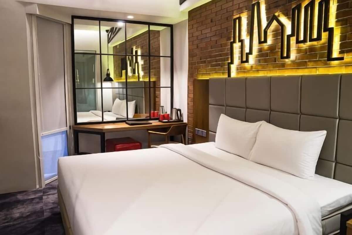 Hotelkamer met een bed, een bureau, een stoel, een spiegel en een bakstenen muur met een geometrische lamp.