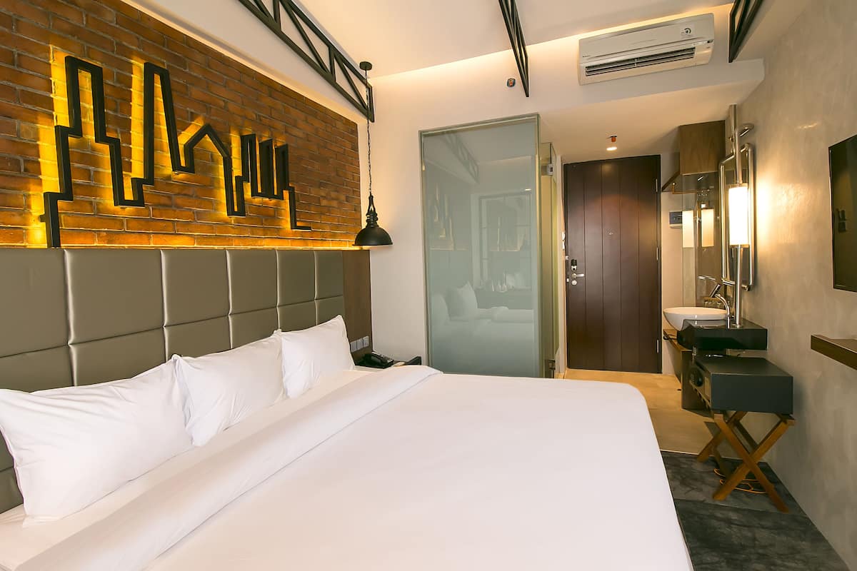 Een moderne hotelkamer met een groot bed, een accentmuur van baksteen, een glazen deur, een badkamer met wastafel en spiegel, en een plafondventilator.