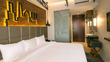 Ein modernes Hotelzimmer mit einem großen Bett, einer Wand aus Ziegelsteinen, einer Glastür, einem Badezimmer mit Waschbecken und Spiegel sowie einem Deckenventilator.
