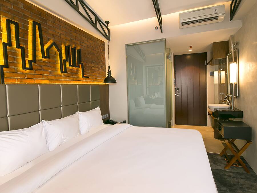 Ein modernes Hotelzimmer mit einem großen Bett, einer Wand aus Ziegelsteinen, einer Glastür, einem Badezimmer mit Waschbecken und Spiegel sowie einem Deckenventilator.