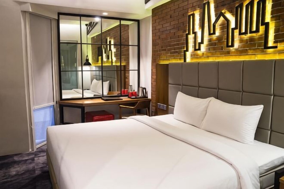 A hotel room with a bed, a desk, a chair, a mirror, and a brick wall with a geometric light fixture.