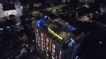 A multi-story hotel with a rooftop terrace, a swimming pool, and a well-lit exterior.