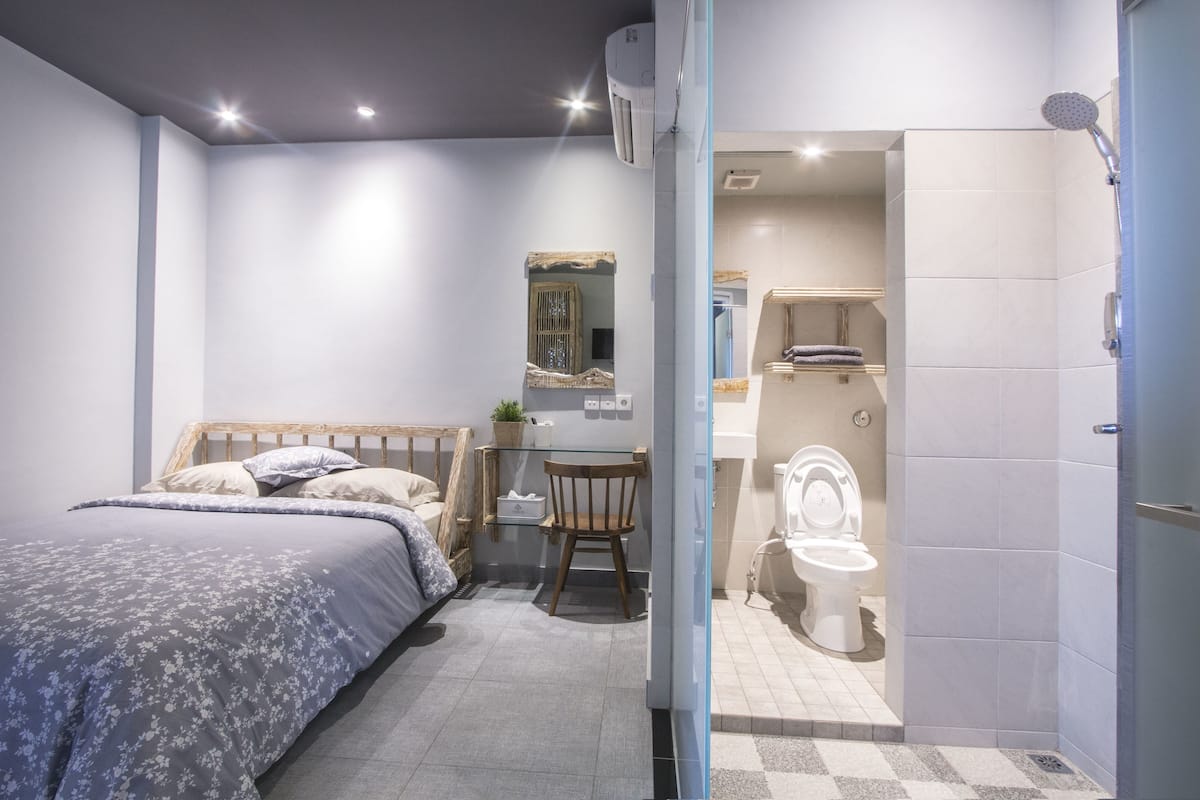 A modern bedroom with a bed, a small table with a chair, a mirror, and a bathroom with a toilet and shower.