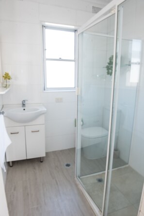 A modern bathroom with a glass shower enclosure, a white sink, and a window. - Lotus Stay Manly - Apartment 29G (Manly)