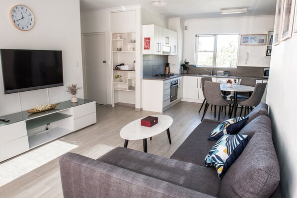 A modern living room with a flat-screen TV, a grey sofa, and a dining area with a table and chairs.