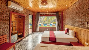 Une pièce avec un plafond en bois, des murs en briques, un lit recouvert d’une couverture rouge, un banc et une vue sur une piscine extérieure.