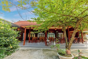 Un área de comedor al aire libre con mesas y sillas de madera, piso de baldosas y un árbol que proporciona sombra.