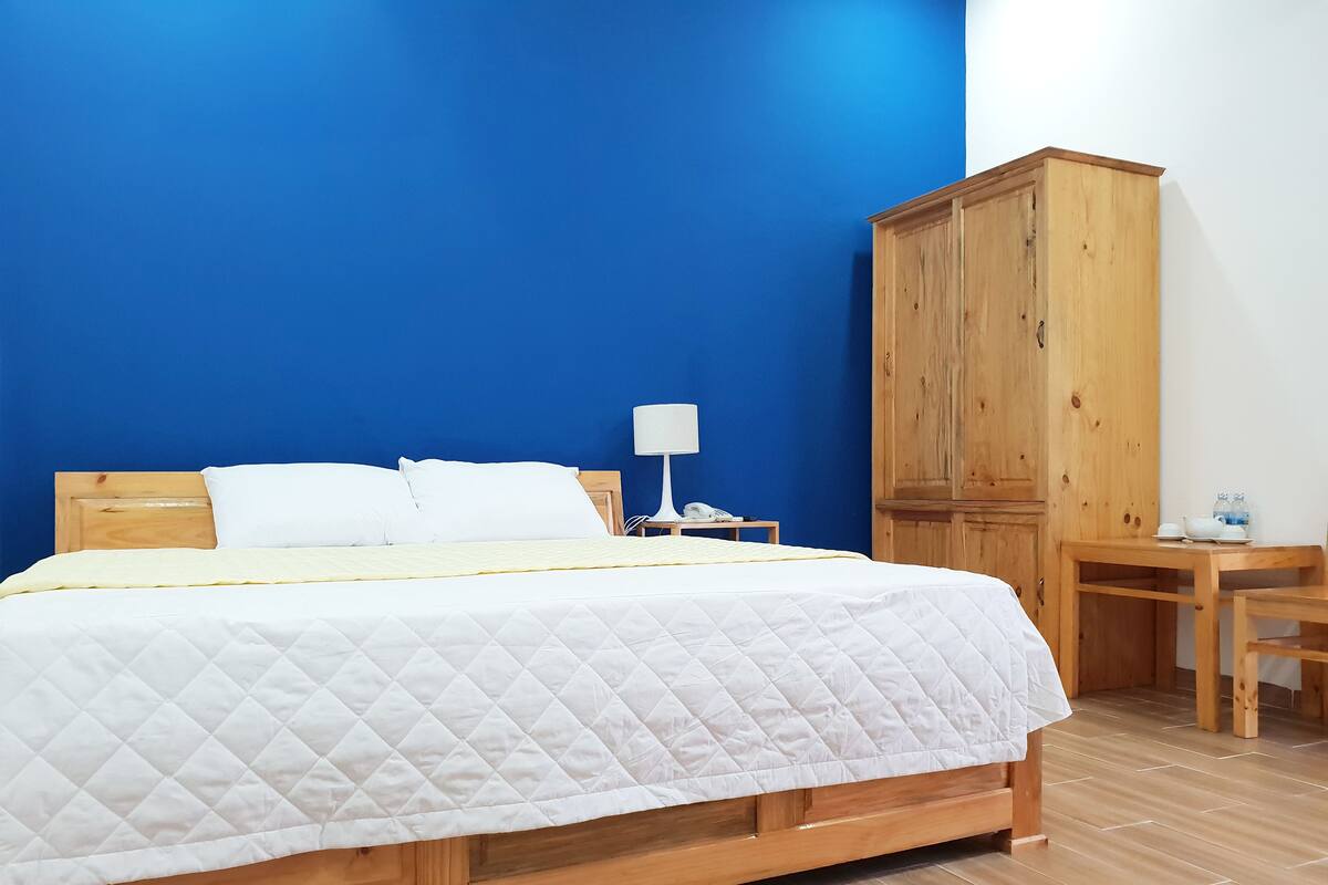 A neatly made bed with a quilted white duvet, a wooden headboard, a bedside table with a lamp, and a wooden wardrobe.